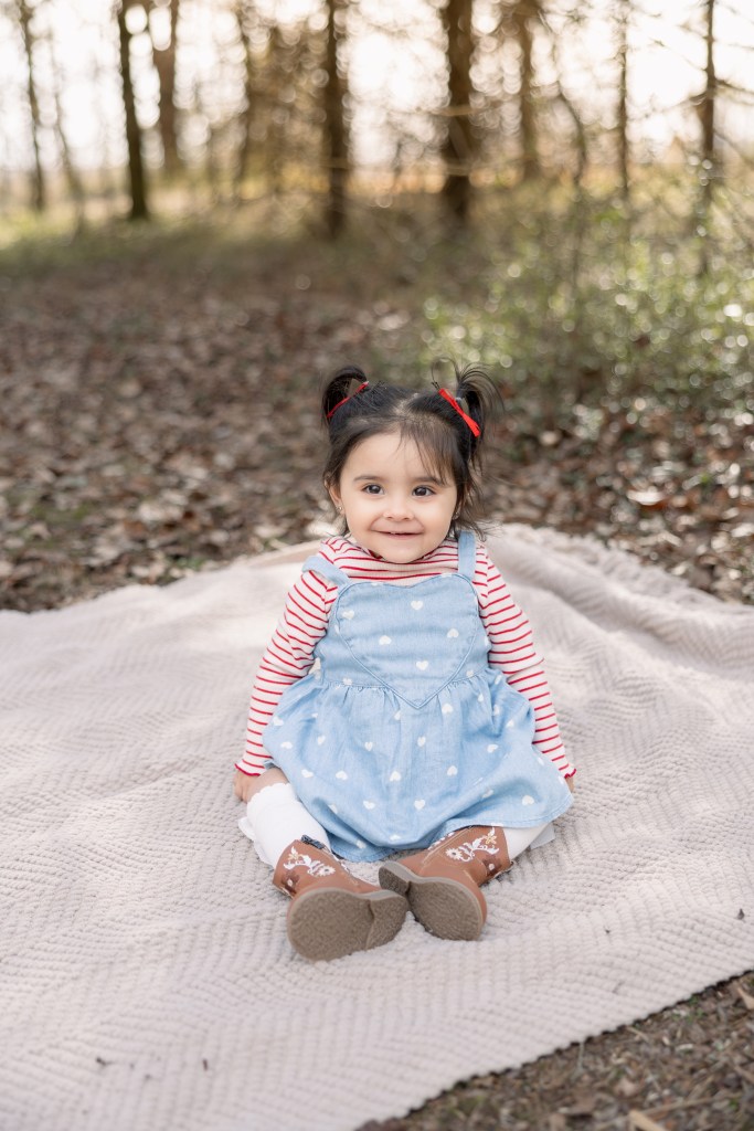 Smiling young girl with dark hair in pigtails, wearing a denim dress with white hearts, a red and white striped shirt, and brown boots, sitting on a beige blanket in a wooded area.